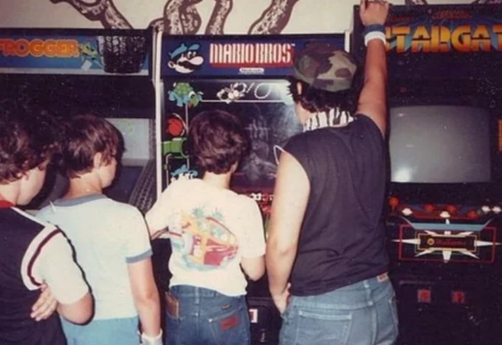 Four kids stand closely together in front of vintage arcade game machines, including Mario Bros., in an arcade. One child is wearing a cap and reaching up, while others watch the screen intently.