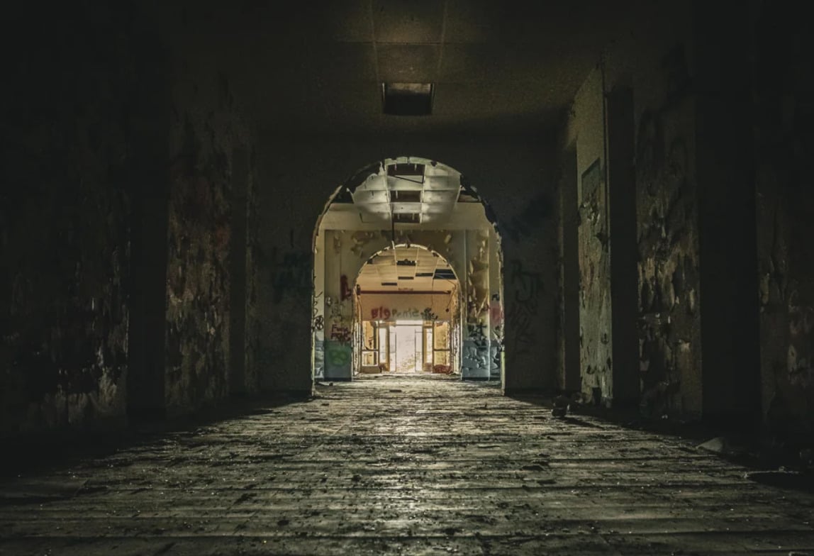 A dark, abandoned hallway with a damaged floor and graffiti-covered walls, lit dimly by distant light coming through a doorway at the end of the corridor.