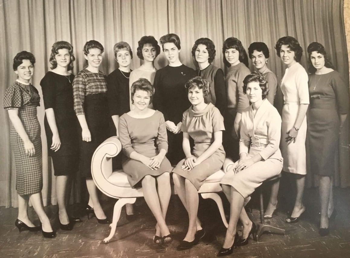 A black-and-white photo of 15 women in formal dresses, posed in two rows; two women sit on a vintage bench while the rest stand behind them, all facing the camera, with a curtain backdrop.
