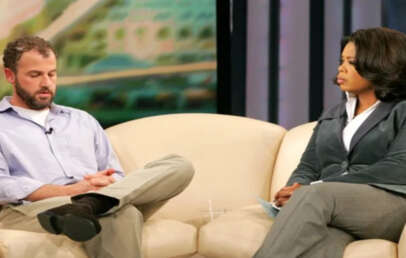A man and a woman sit on a beige couch during a talk show interview. The man has a beard, wears a light blue shirt, and looks down with his hands clasped. The woman listens attentively in a gray suit.
