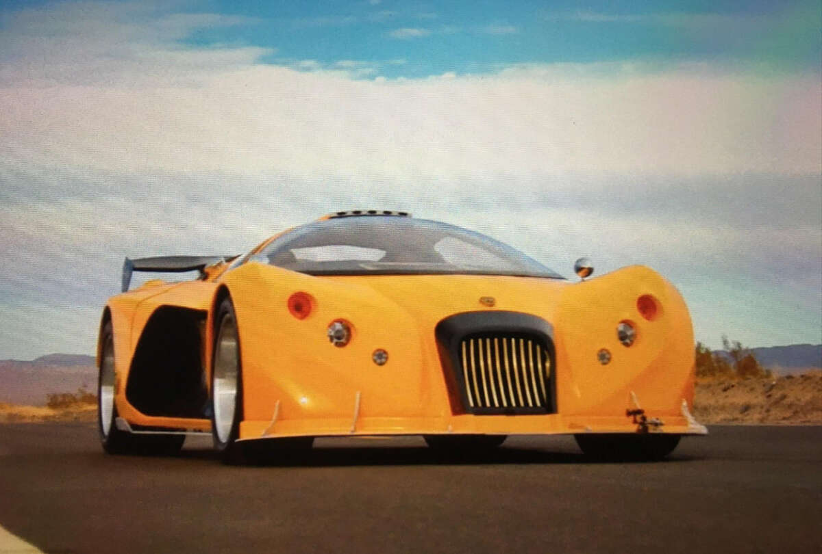 A low-angle view of a bright yellow sports car with a unique front grille and round headlights, driving on an open road with a blue sky and desert landscape in the background.