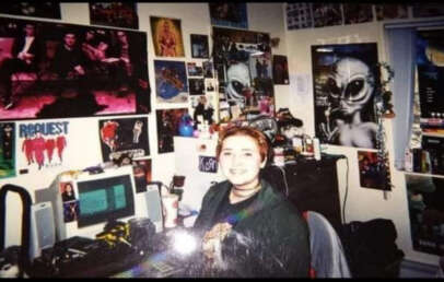 A person with short hair smiles while sitting at a cluttered desk with a computer in a bedroom. The walls are covered with posters, including an alien, “The Matrix,” and various other movies and bands.