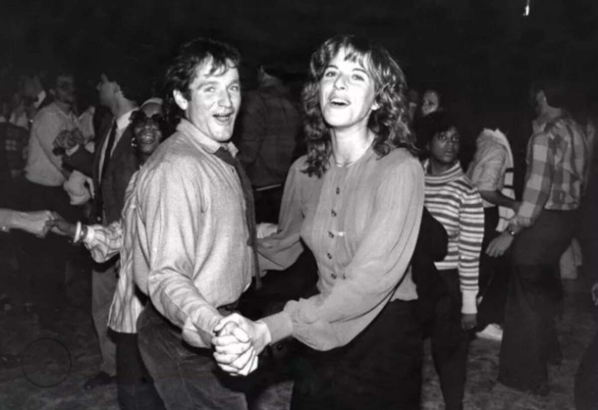 A man and woman smile and hold hands while dancing at a lively indoor event, surrounded by other people enjoying themselves. The image is in black and white.
