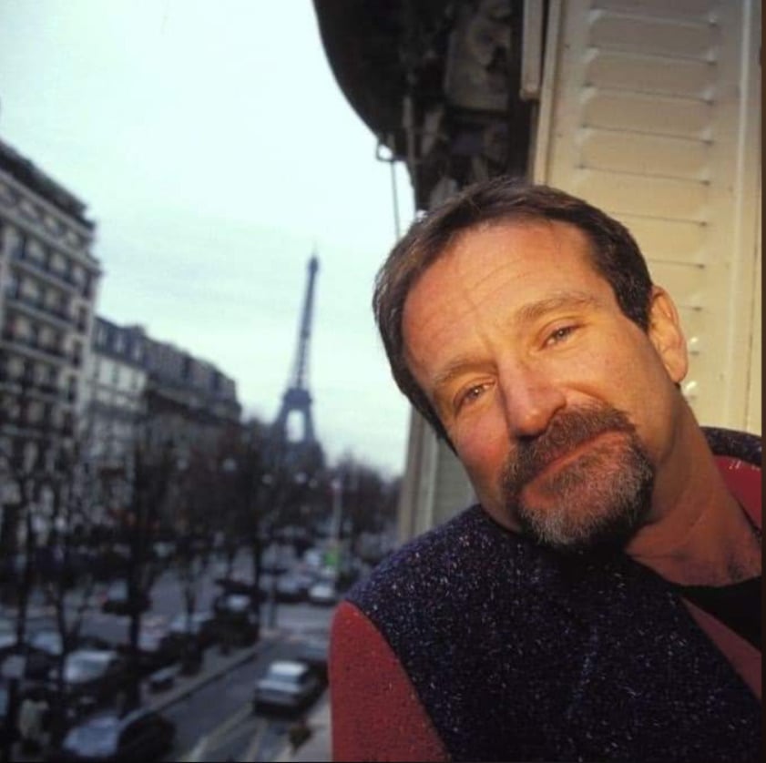 A man with short hair and a beard leans out of a window, smiling gently. The Eiffel Tower and Parisian cityscape are visible in the background.
