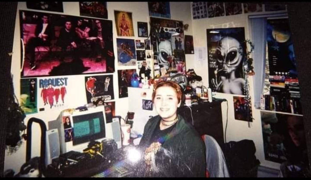 A person with short hair smiles at a desk in a cluttered room decorated with posters of movies, aliens, and bands, with a computer and various objects on the desk and shelves.