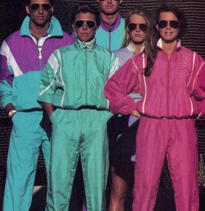 Five people wearing colorful, retro 1980s tracksuits and sunglasses stand confidently together. The tracksuits are teal, purple, and pink, and the group poses against a dark background.