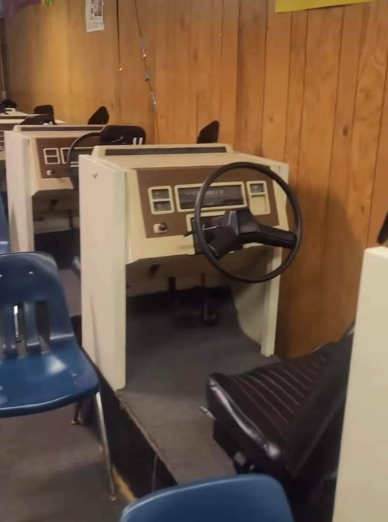 A row of vintage driving simulators with steering wheels and car seats is set up against a wood-paneled wall, with blue plastic chairs placed nearby.