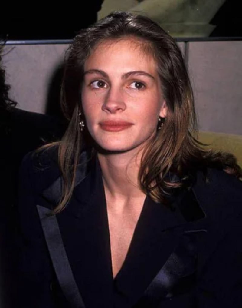 A young woman with light skin, long brown hair, and wearing a dark blazer sits indoors, looking slightly up and to the side with a neutral expression.