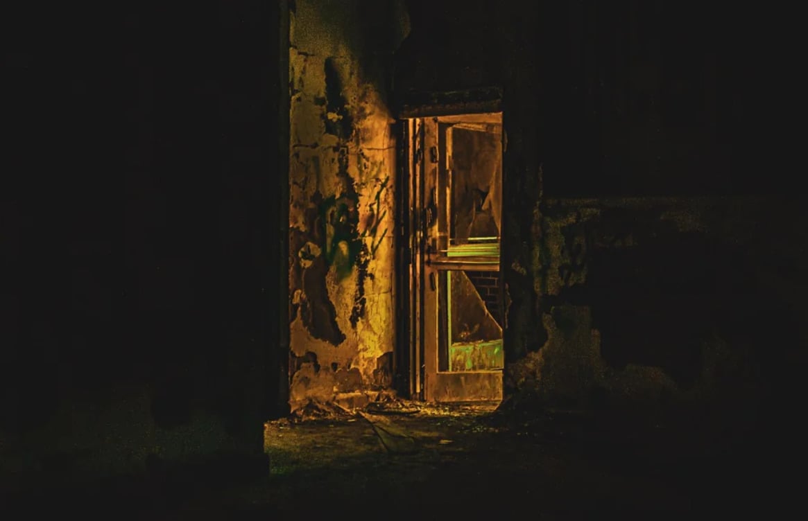 A dimly lit, crumbling doorway glows with eerie yellow-green light, partially illuminating peeling walls and debris in an otherwise dark, abandoned building.