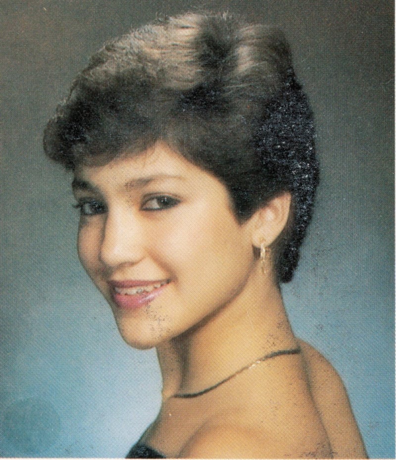 A young woman with short, dark hair smiles at the camera over her shoulder. She is wearing a black top, small hoop earrings, and a thin necklace. The background is a soft, gradient blue.