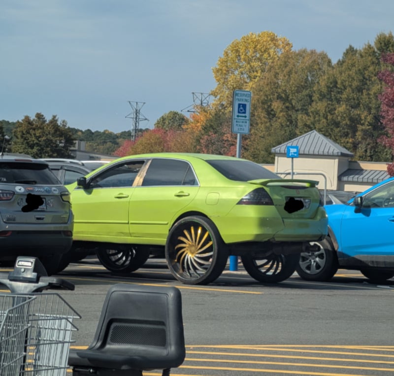A bright green sedan with extremely large, shiny custom rims is parked in a lot near a handicap parking sign. Shopping carts and other cars are nearby, with autumn trees in the background.