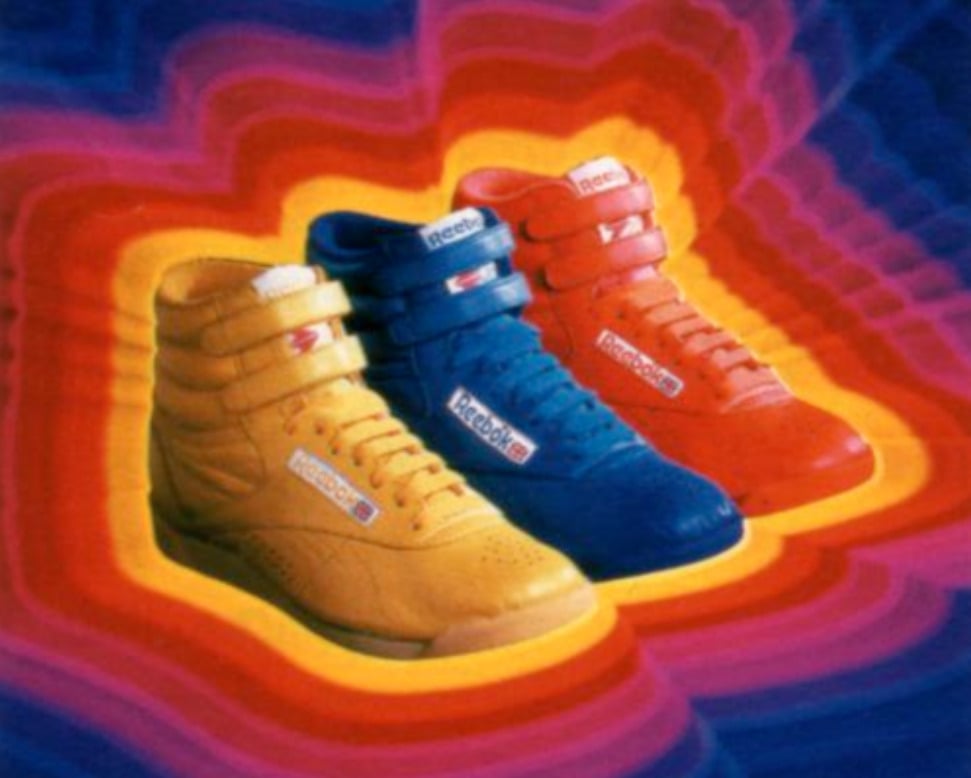 Three high-top Reebok sneakers in yellow, blue, and red are displayed side by side against a vibrant, layered background with wavy rings of pink, purple, orange, and red.
