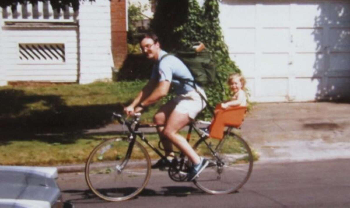 A man rides a bicycle on a suburban street with a small child smiling in a child seat attached to the back. The man wears shorts and glasses, and the background shows a white house and garage.