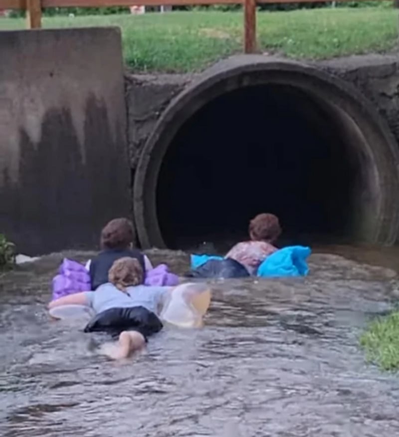Three people on floaties paddle toward the opening of a large concrete drainage pipe in shallow, flowing water, with grass and a wooden fence visible above the pipe.