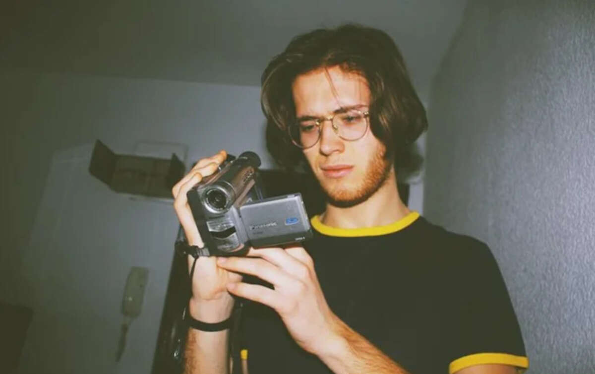 A young person with glasses and shoulder-length hair, wearing a black t-shirt with yellow trim, looks closely at a handheld video camera in a dimly lit room.