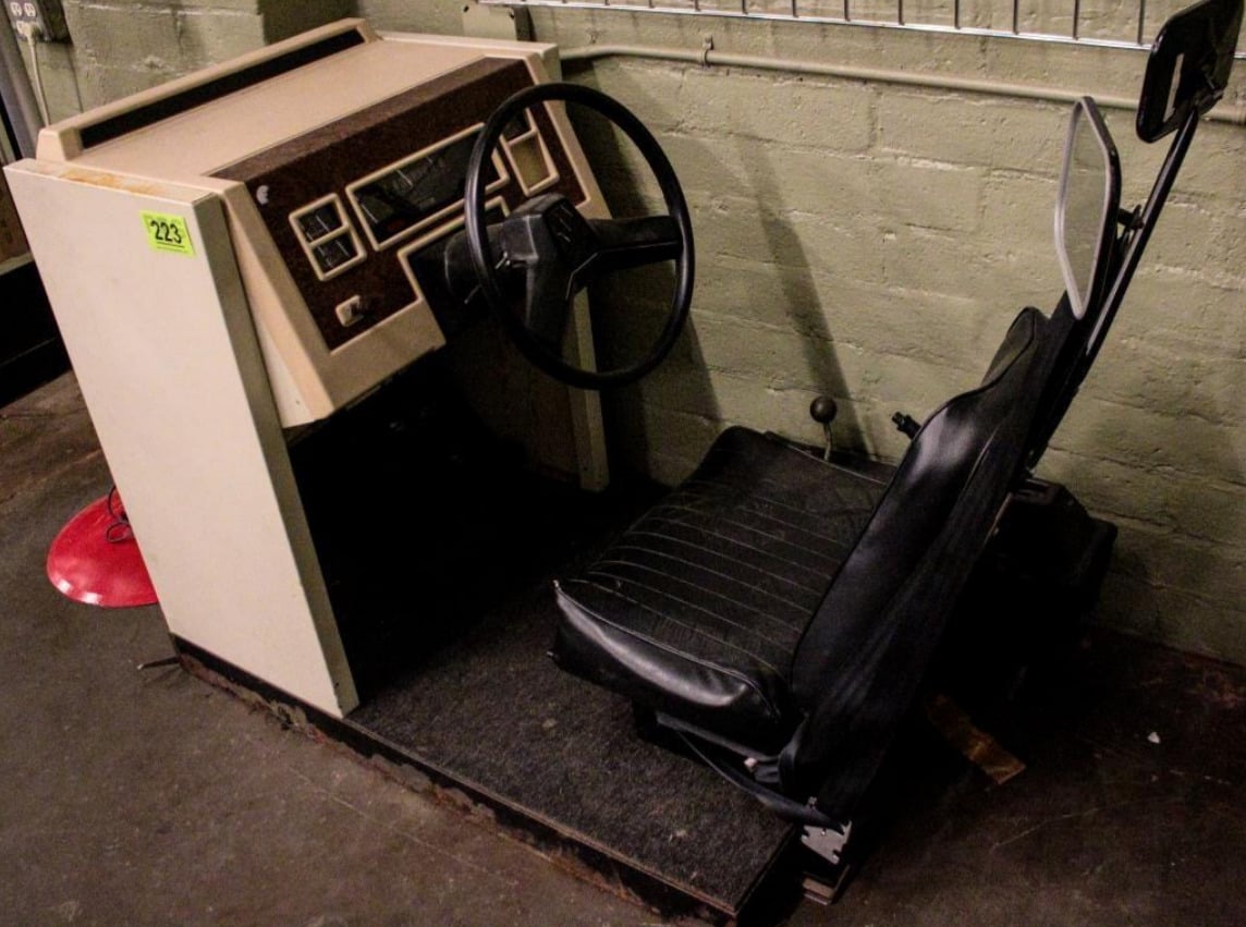 A vintage driving simulator features a steering wheel, dashboard with controls, and a black seat, all mounted on a platform against a brick wall.