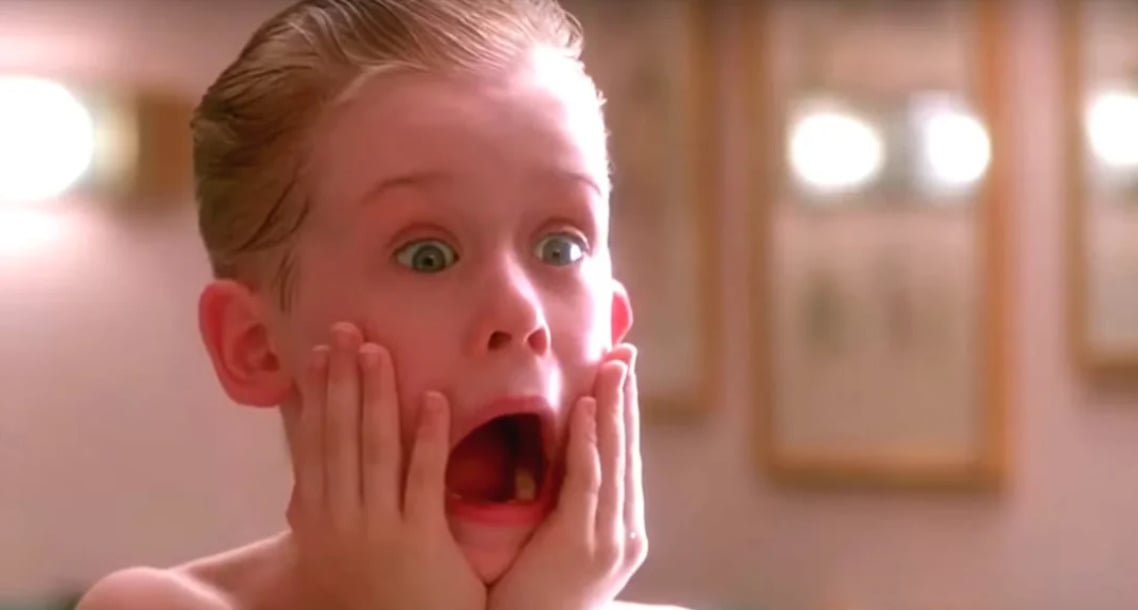 A young boy with slicked-back hair holds his hands to his cheeks and looks shocked, mouth open wide and eyes wide, in a well-lit room with blurry picture frames in the background.