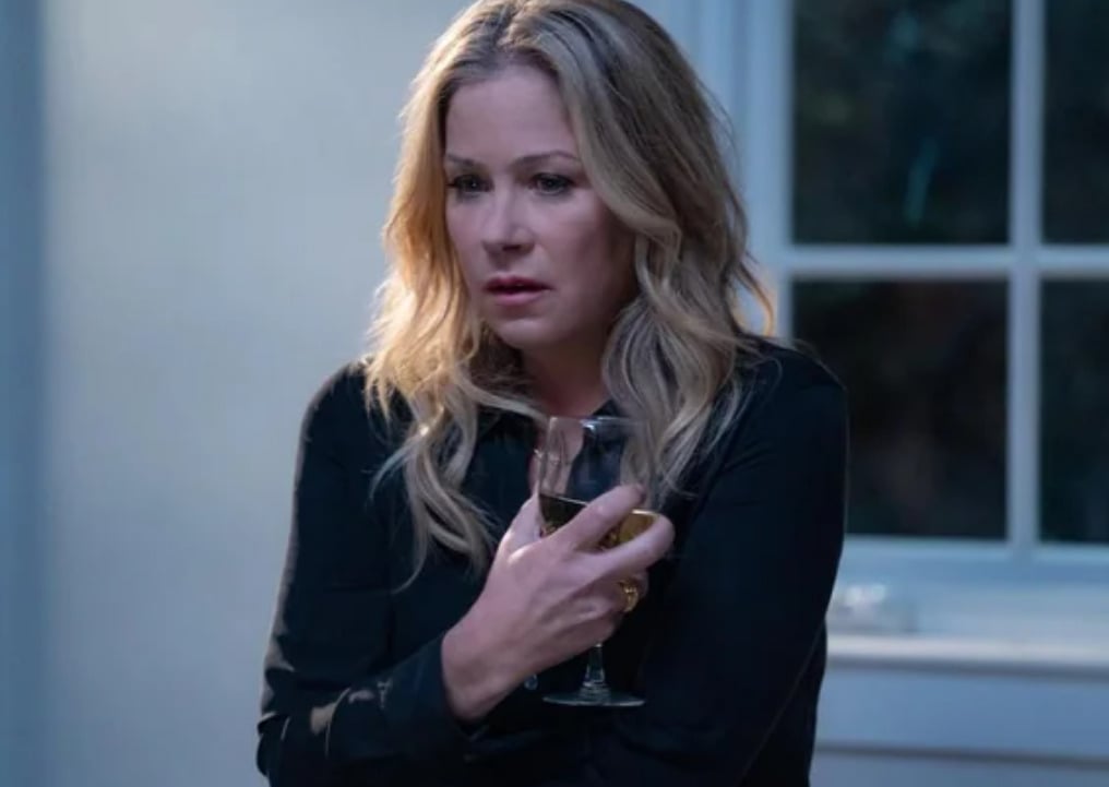 A woman with long blonde hair wearing a dark shirt holds a glass of white wine and looks troubled, standing indoors near a window.