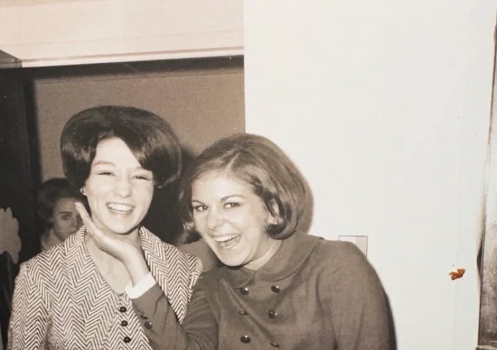 Two women in vintage clothing smile and laugh together indoors; one playfully holds the other’s chin. The photo appears to be from the 1960s and is in black and white. Another person is visible in the background.