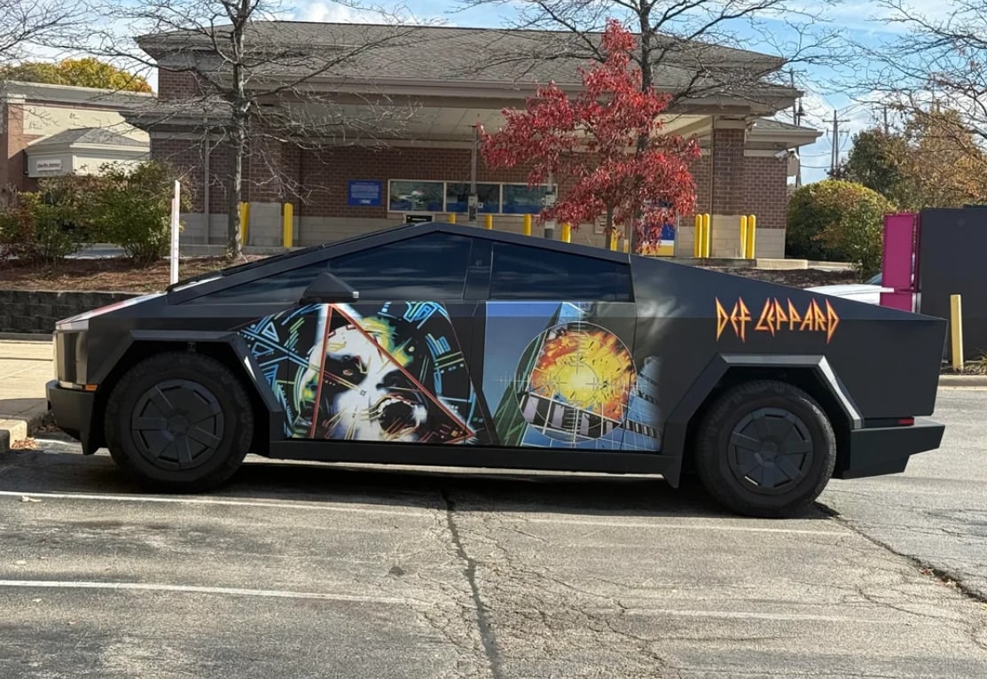 A matte black Tesla Cybertruck is parked in a lot, decorated with vibrant Def Leppard album artwork and the band's name painted on the side. Trees with autumn leaves and a brick building are in the background.