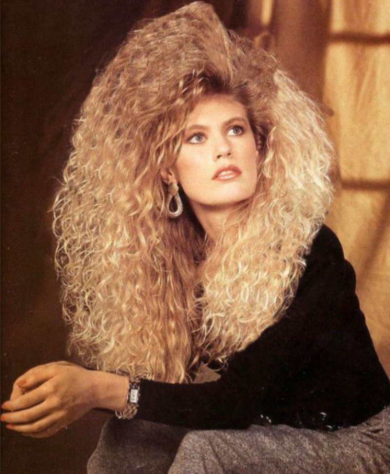 A woman with voluminous, curly blond hair styled in a dramatic 1980s fashion, wearing hoop earrings, a black top, and a watch, looks thoughtfully to the side against a warm-toned background.