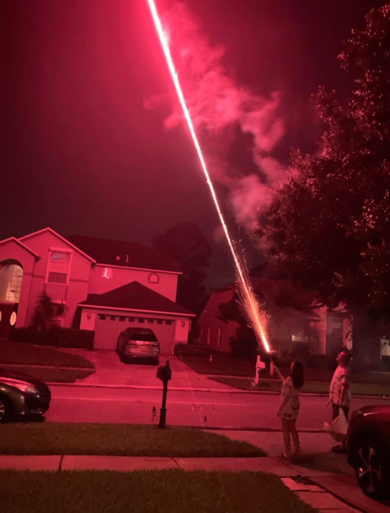 A person standing on a suburban street at night aims a bright red firework or flare into the sky, creating a vivid streak of light with smoke. Two houses and several cars are visible in the background.