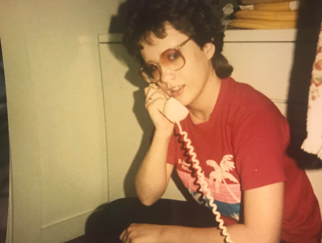 A person with short hair and glasses sits on the floor wearing a red T-shirt with palm trees, holding a corded telephone to their ear and looking slightly to the side.