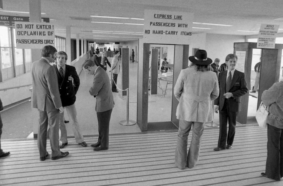 Black and white photo of an airport security checkpoint in the 1970s; several men in suits and hats talk near signs that direct deplaning passengers and those with no hand-carry items to different lines.