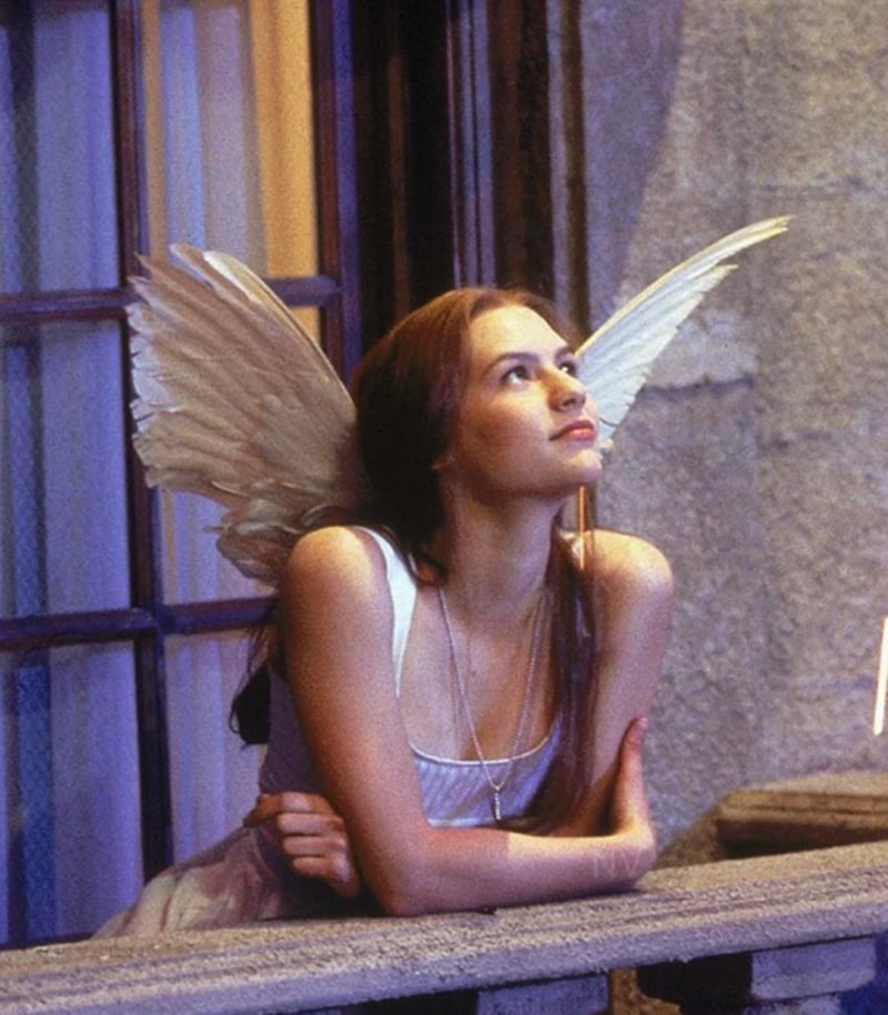 A woman with long brown hair, wearing delicate white fairy wings and a white dress, leans on a stone balcony railing and gazes upward thoughtfully, with a soft light illuminating her face.