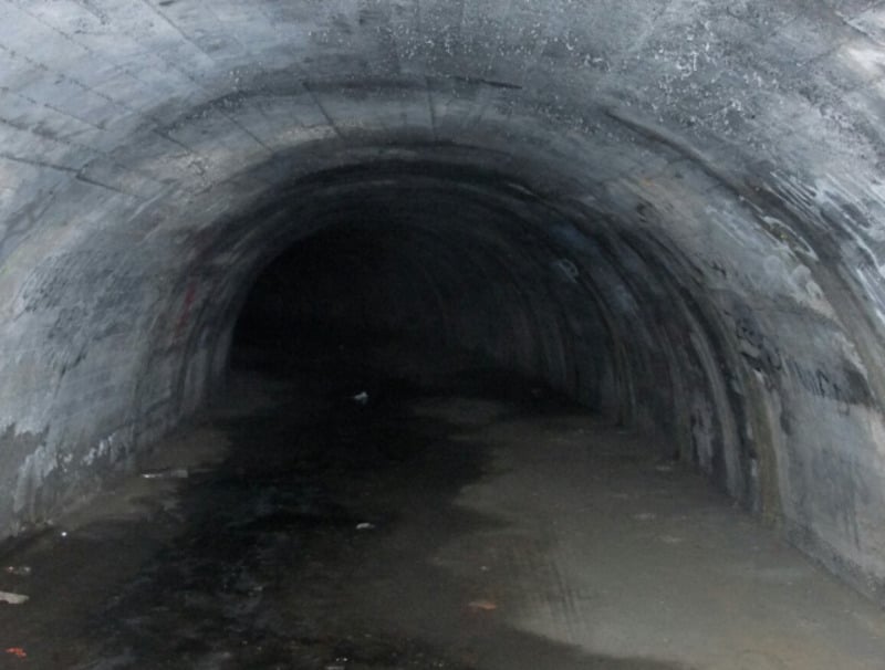 A dark, empty concrete tunnel with a curved ceiling, wet floor, and dim lighting. The tunnel extends into darkness, with water stains and debris visible along the sides.
