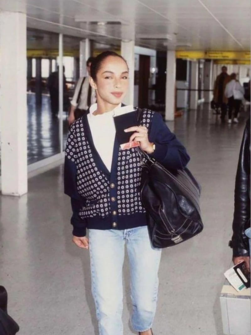 A woman wearing a patterned navy cardigan, white shirt, and light blue jeans stands in an airport terminal, holding a passport and a large black bag, smiling at the camera.
