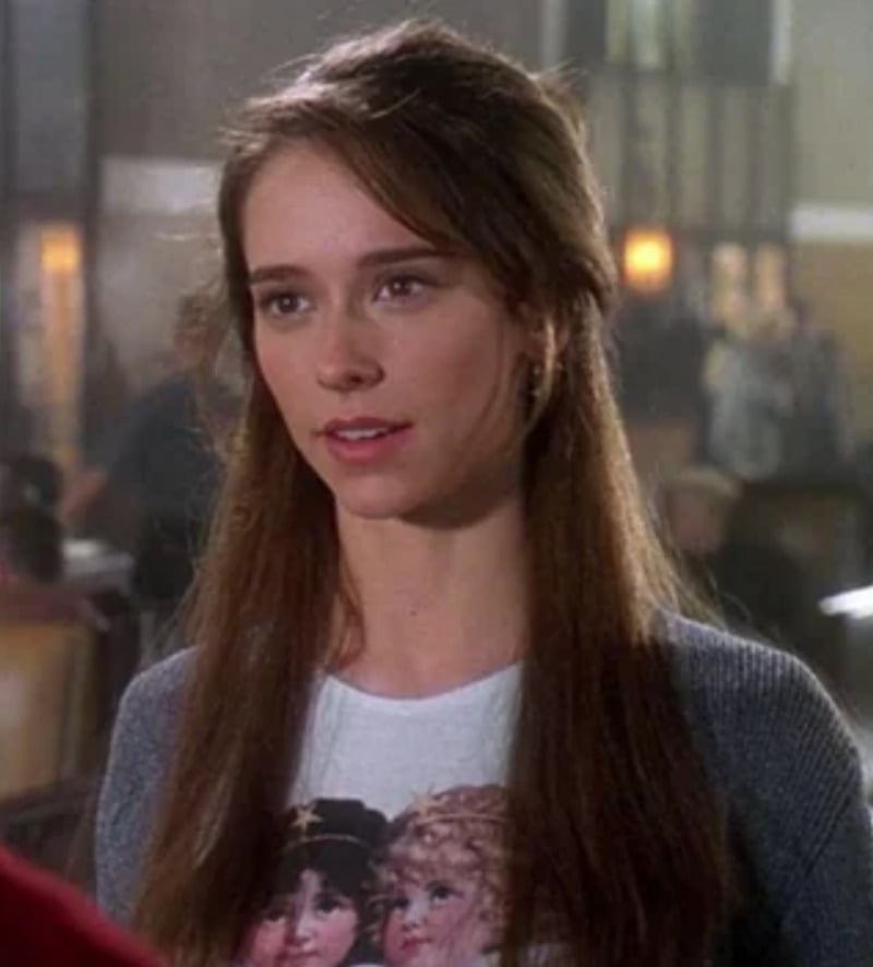 A young woman with long brown hair, wearing a white shirt with a print of two dolls and a gray cardigan, stands indoors with a soft, neutral expression. The background is blurred, showing a busy, well-lit room.