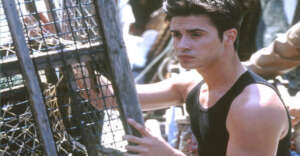 A young man with short dark hair wearing a sleeveless black shirt stands outdoors, holding onto a wooden structure with wire mesh, possibly a lobster trap, while looking off to the side.