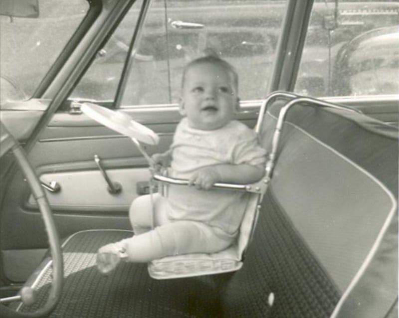 A black and white photo of a baby sitting in an old-fashioned car seat attached to the front bench seat of a vintage car, holding a toy steering wheel and looking up, with the car door and window visible.