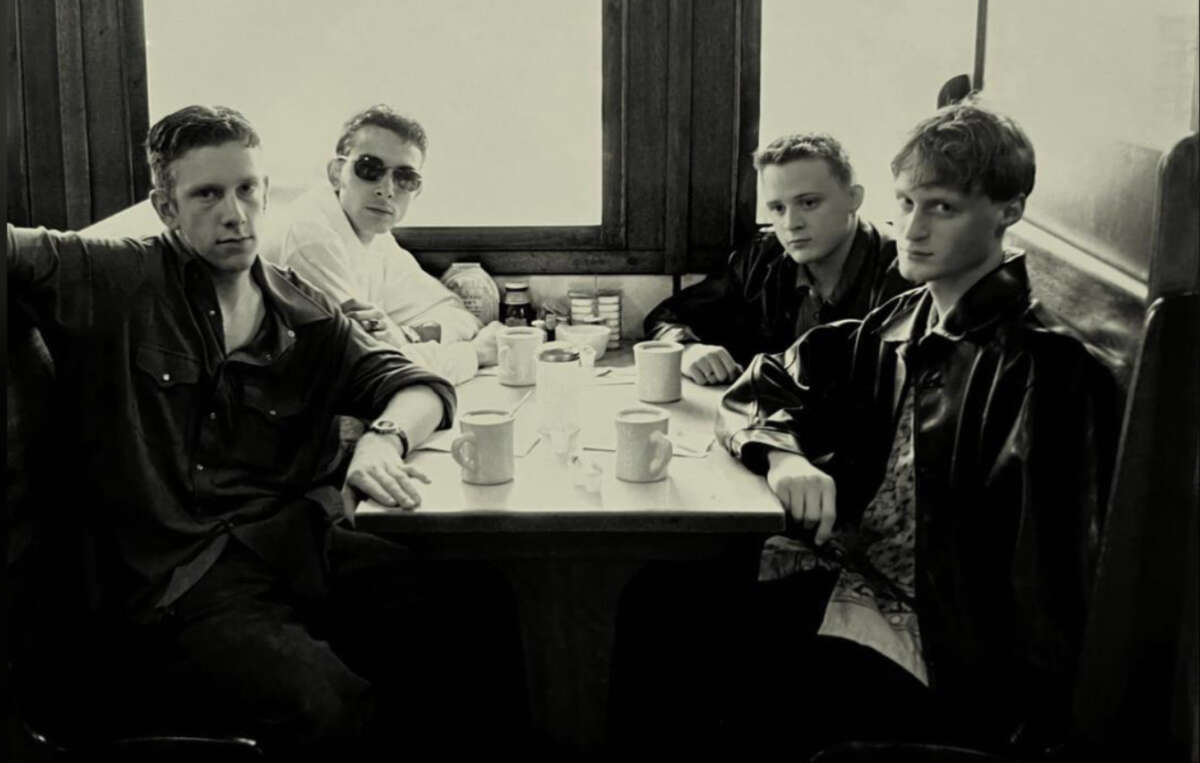 Four young men sit around a diner table with coffee mugs and condiments, looking at the camera. The photo is in black and white, with light streaming through the windows behind them.