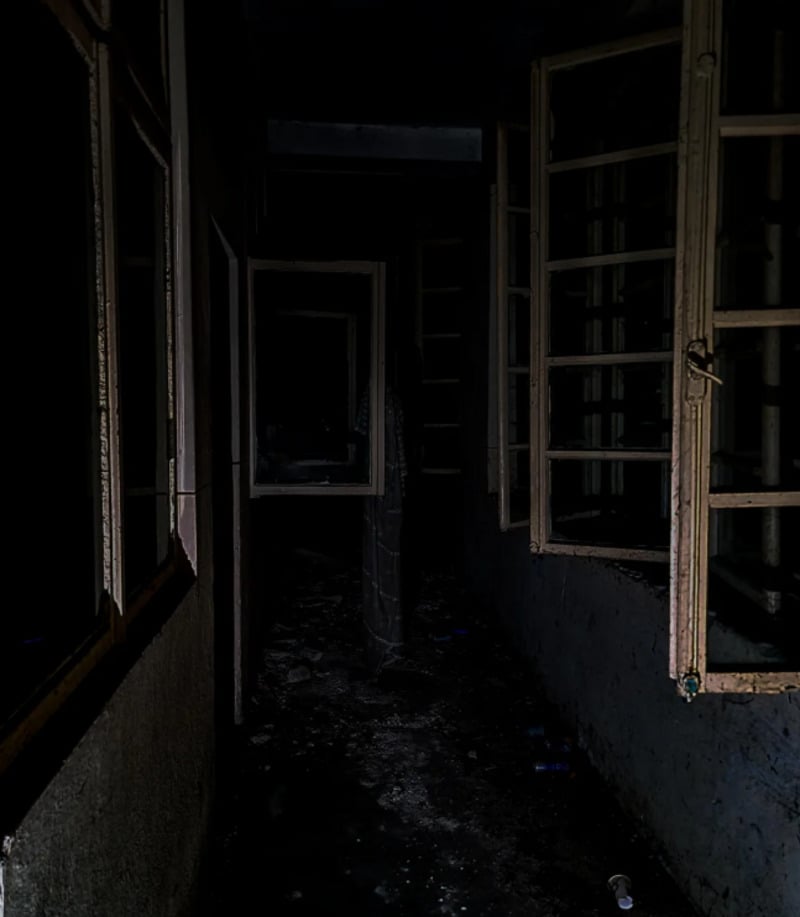A dark, narrow hallway with open windows on the right side, showing peeling paint and grime. The corridor appears abandoned, with scattered debris and faint light highlighting the eerie, shadowy atmosphere.