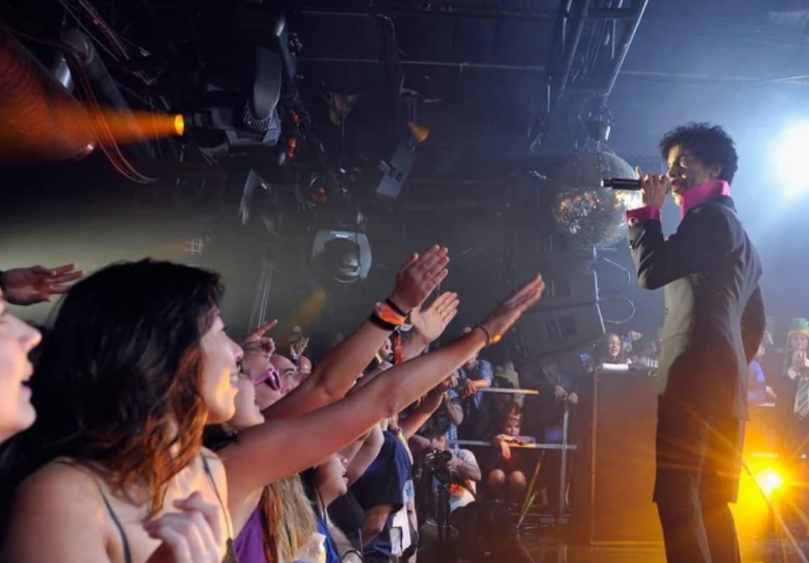 A singer in a dark, stylish outfit performs on stage with a mic, facing an enthusiastic crowd with raised hands. Stage lights and a disco ball create a vibrant, energetic concert atmosphere.