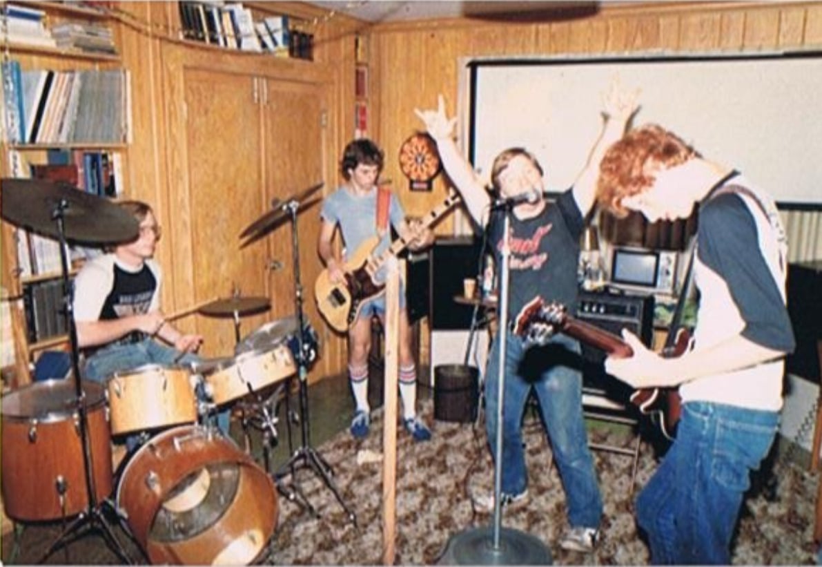 Four teenage boys play in a band in a wood-paneled room; one plays drums, another bass, one sings passionately at the microphone, and another plays guitar. The room has bookshelves, records, and a dartboard.