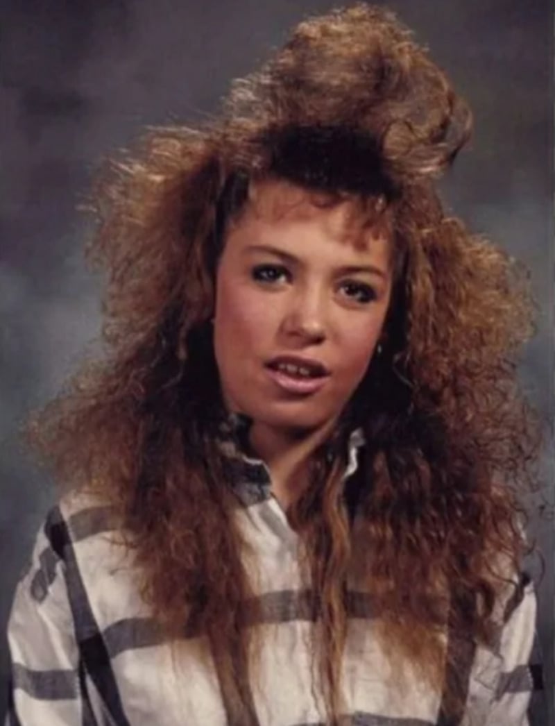 A young woman with voluminous, teased curly hair wears a striped button-up shirt and poses in front of a gray studio backdrop, giving a slight smile.