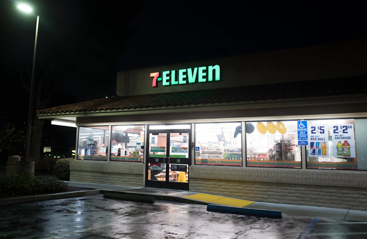 A 7-Eleven convenience store at night, brightly lit inside, with a visible entrance door, large windows displaying products, and wet pavement in the nearly empty parking lot outside.