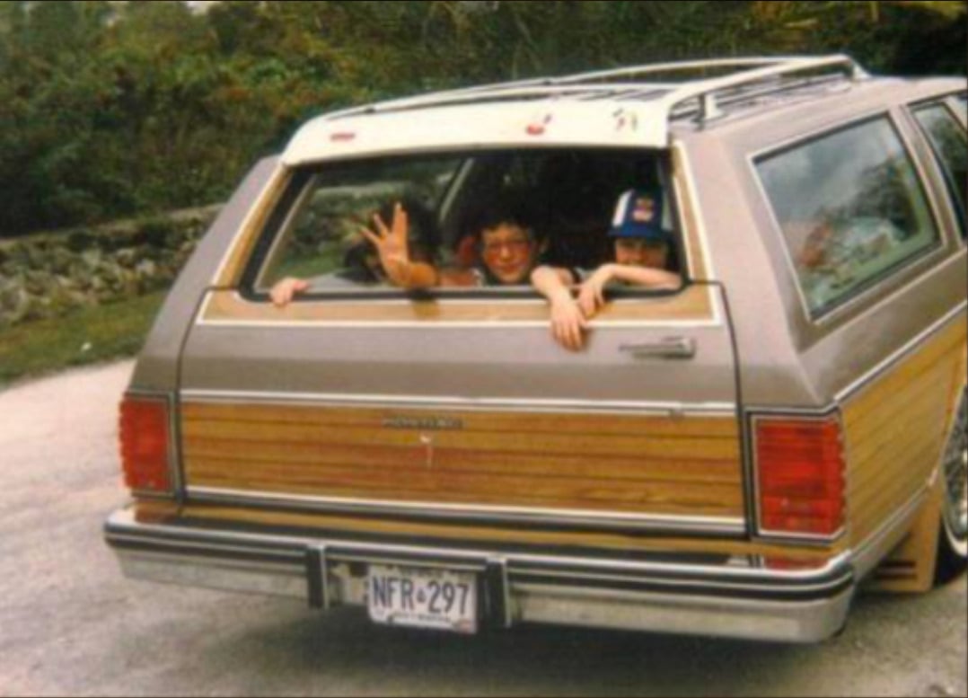 Two children wave and smile from the open rear window of a vintage brown station wagon with wood paneling, parked on a road with greenery and a stone wall in the background.