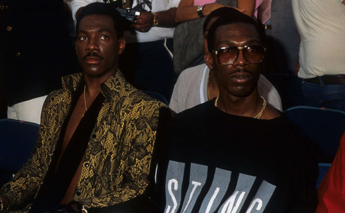 Two men sit side by side in a crowd. The man on the left wears a gold snakeskin-patterned jacket, while the man on the right wears sunglasses and a black shirt with large "STING" lettering. Other people are visible behind them.