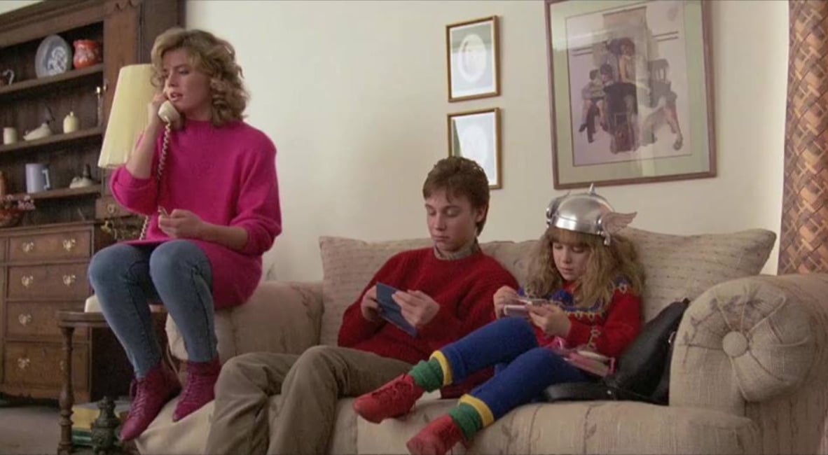 A woman in a pink sweater talks on a corded phone while sitting on a couch arm. Next to her, a teenage boy and a young girl in a winged helmet play with handheld devices, all in a cozy living room.