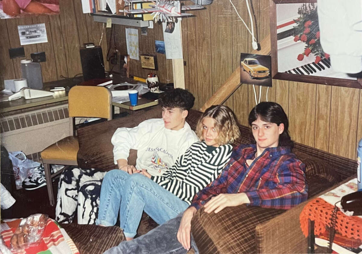 Three teenagers sit closely together on a brown couch in a wood-paneled room decorated with posters, a desk, and various items. Two of them wear striped shirts, and one wears a plaid shirt. The atmosphere appears relaxed and casual.
