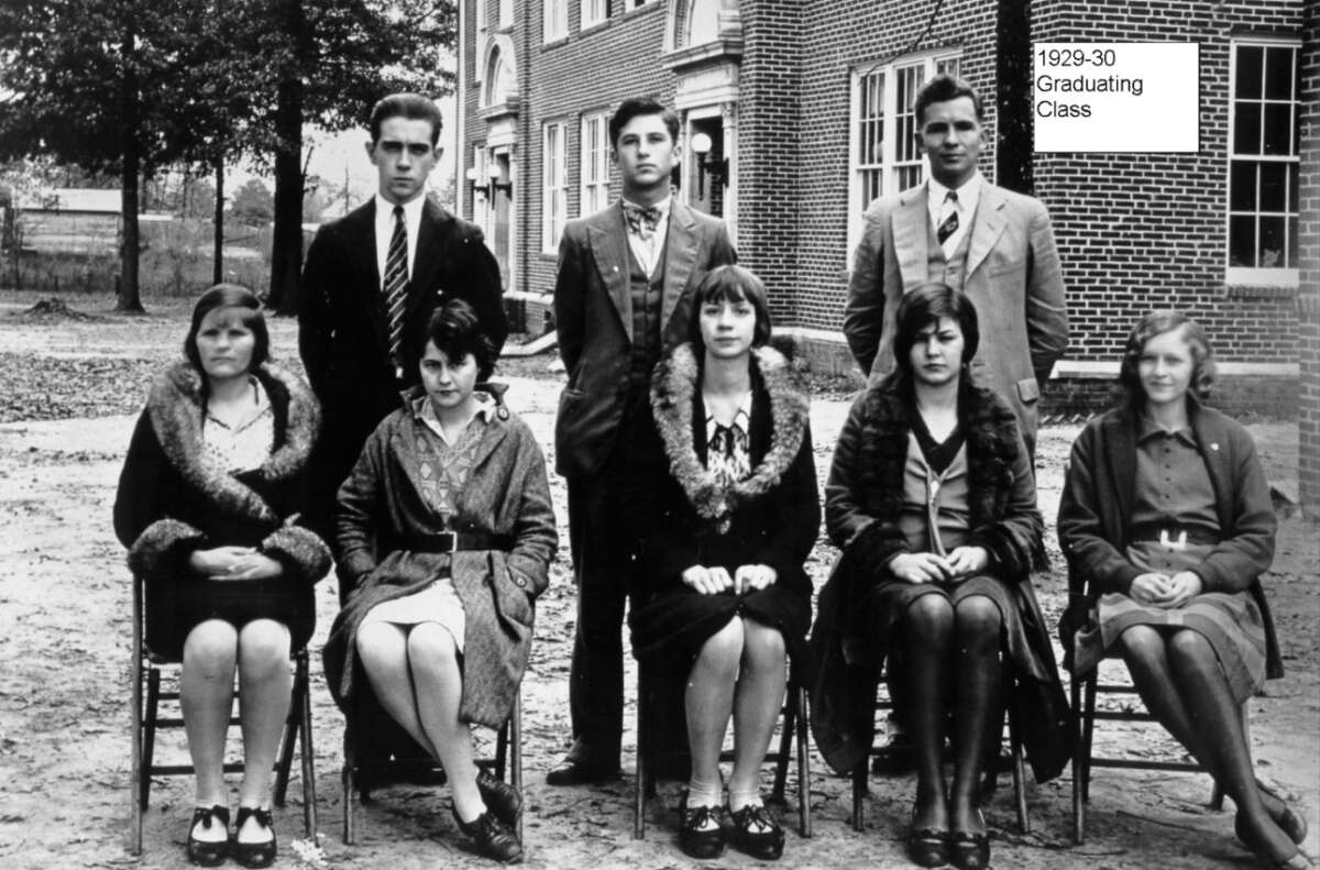 Black and white photo of the 1929-30 graduating class. Four men stand behind four women seated in front, all in formal attire, outside a brick building with trees in the background.