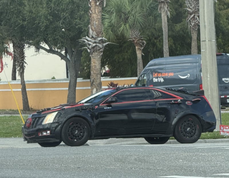A black car with red accents and custom modifications is stopped at an intersection. Palm trees and an Amazon delivery van are visible in the background.