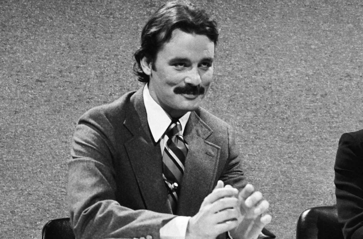 A man with a mustache wearing a suit and striped tie sits at a table, smiling and holding his hands together in front of a plain, textured background. The image is in black and white.