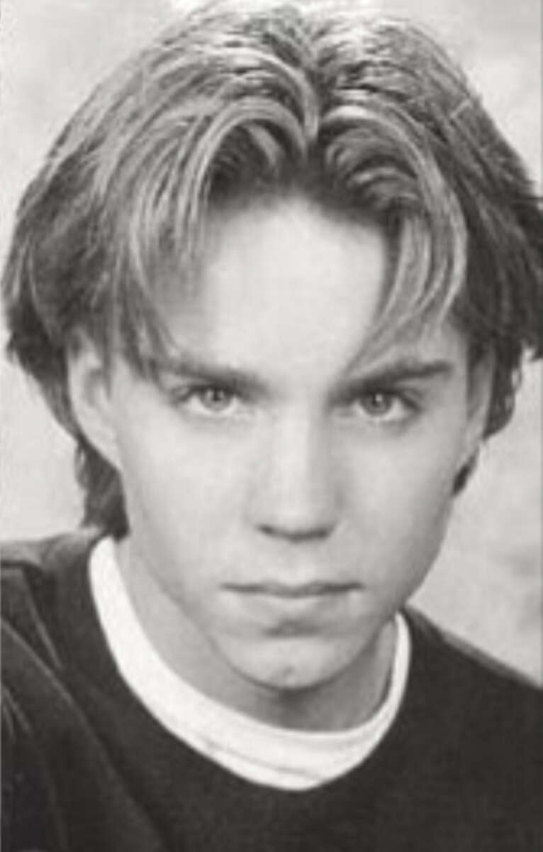 Black and white portrait of a young person with light, center-parted hair, looking directly at the camera with a serious expression, wearing a dark shirt over a light collar.