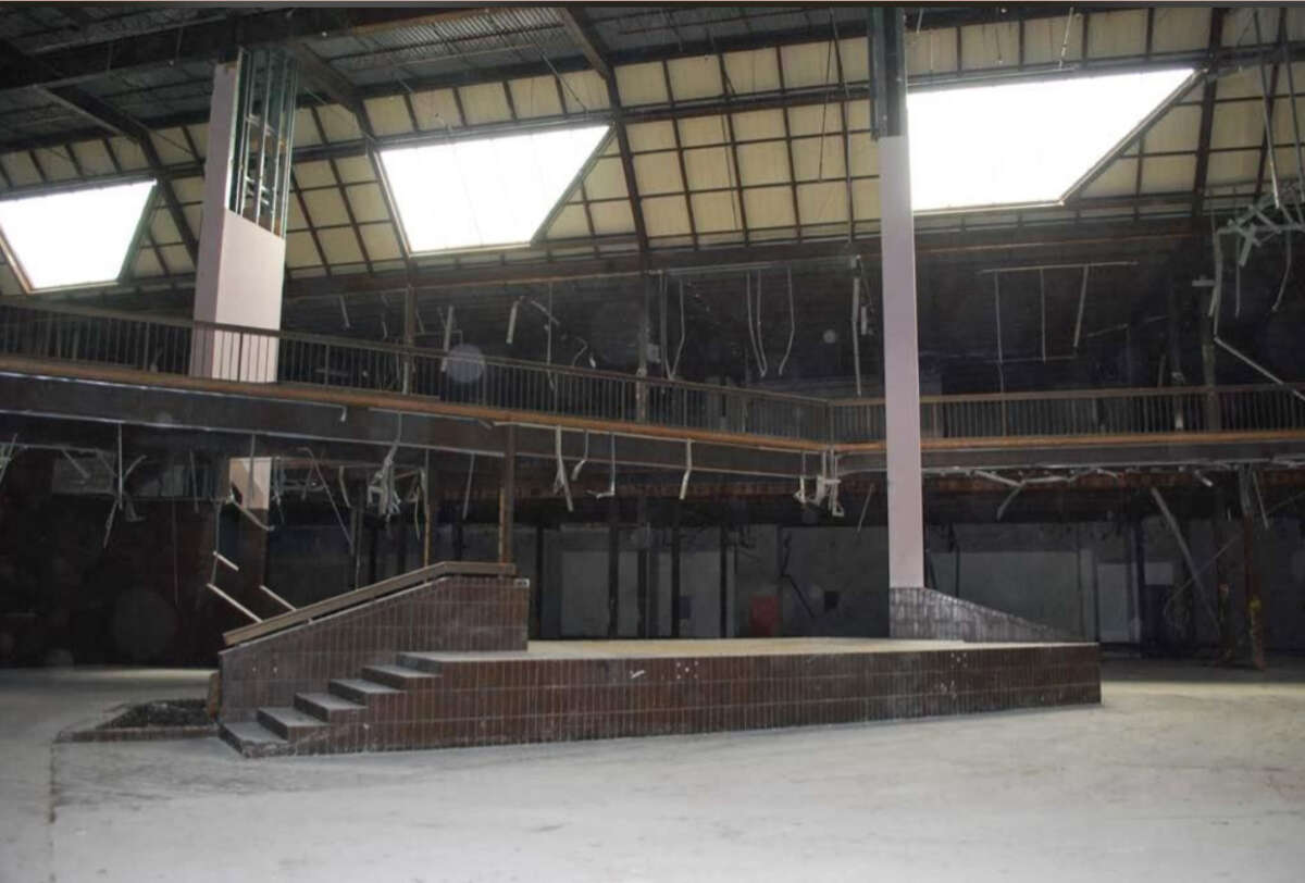 An empty, abandoned industrial building with a stage-like platform, brick stairs, high ceiling, exposed beams, and hanging debris. Light filters in through large triangular skylights.