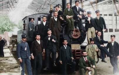 A group of men in late 19th-century clothing pose around and on a large steam locomotive inside a train shed, with some men in uniform and others in suits, standing or sitting casually on the engine.